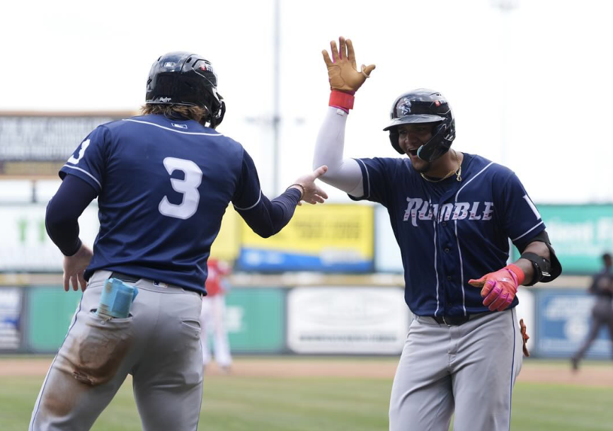 Two baseball players in navy blue uniforms celebrate on the field during a sports game, giving each other a high-five. One players jersey reads Rumble and both wear helmets and gloves. The background shows a baseball field and advertisements.